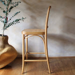 Wooden bar stool next to a woven vase with greenery on a wooden floor.