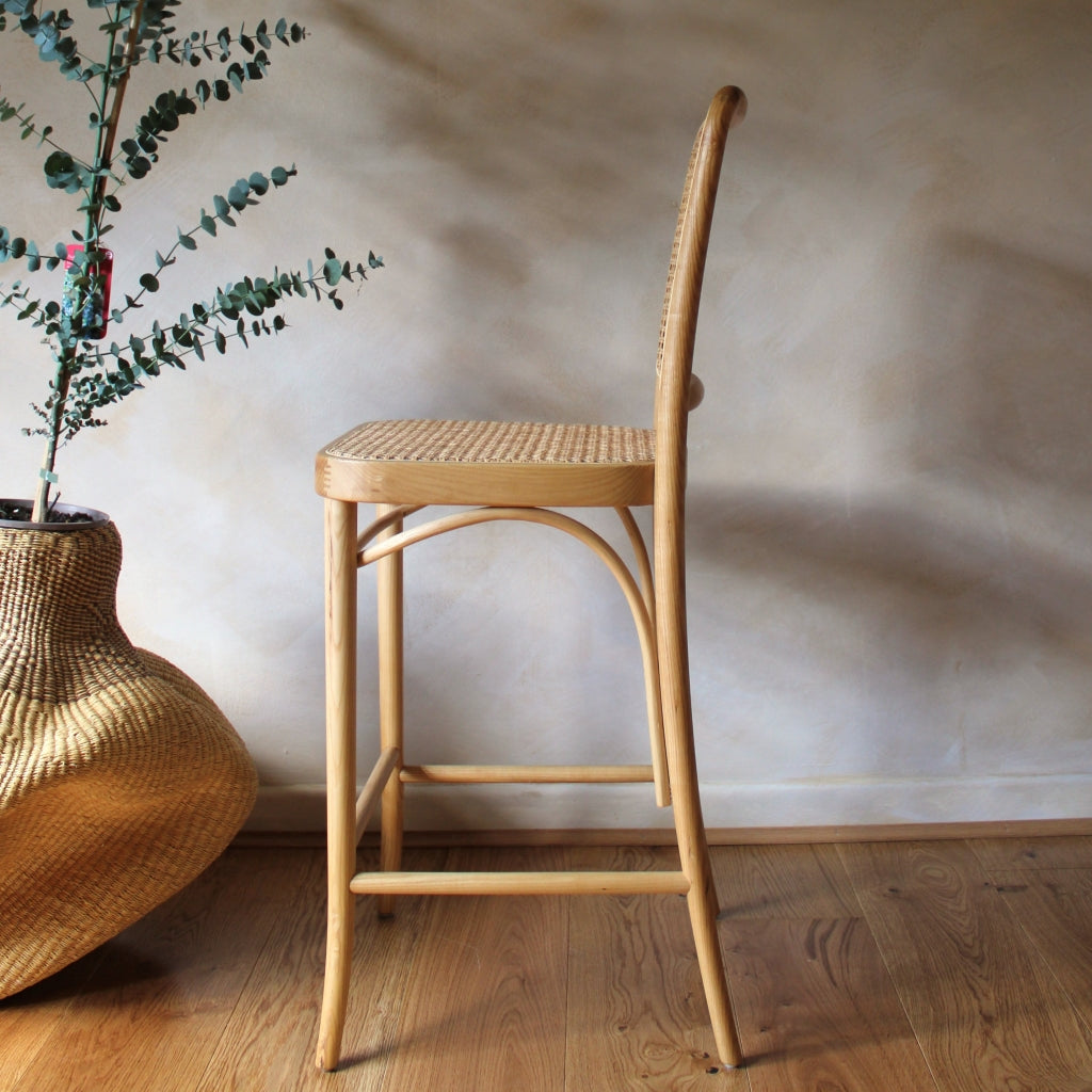 Wooden bar stool next to a woven vase with greenery on a wooden floor.